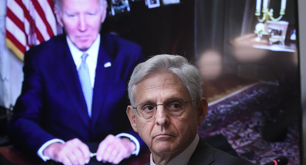 Attorney General Merrick Garland in a suit in front of a screen showing President Joe Biden in a blue suit.