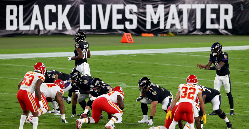 NFL teams Chiefs and Raven wear black, red and white uniforms, squatting in position for the next play, while a BLM sign is displayed behind.