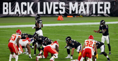 NFL teams Chiefs and Raven wear black, red and white uniforms, squatting in position for the next play, while a BLM sign is displayed behind.