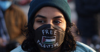 A girl wear a "Free Palestine" black mask, with a grey beanie on. She is looking off into the distance.