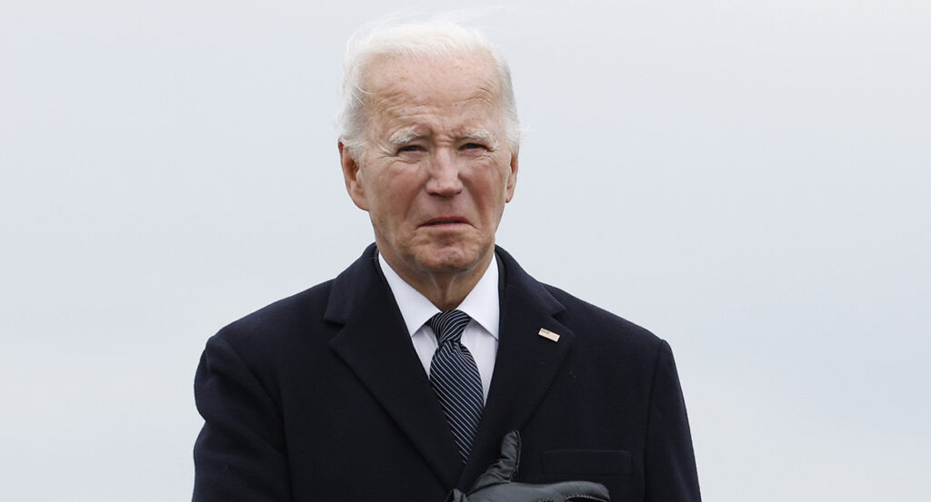 President Joe Biden grimaces with a blank stare in a black suit, holding his hand against his heart.