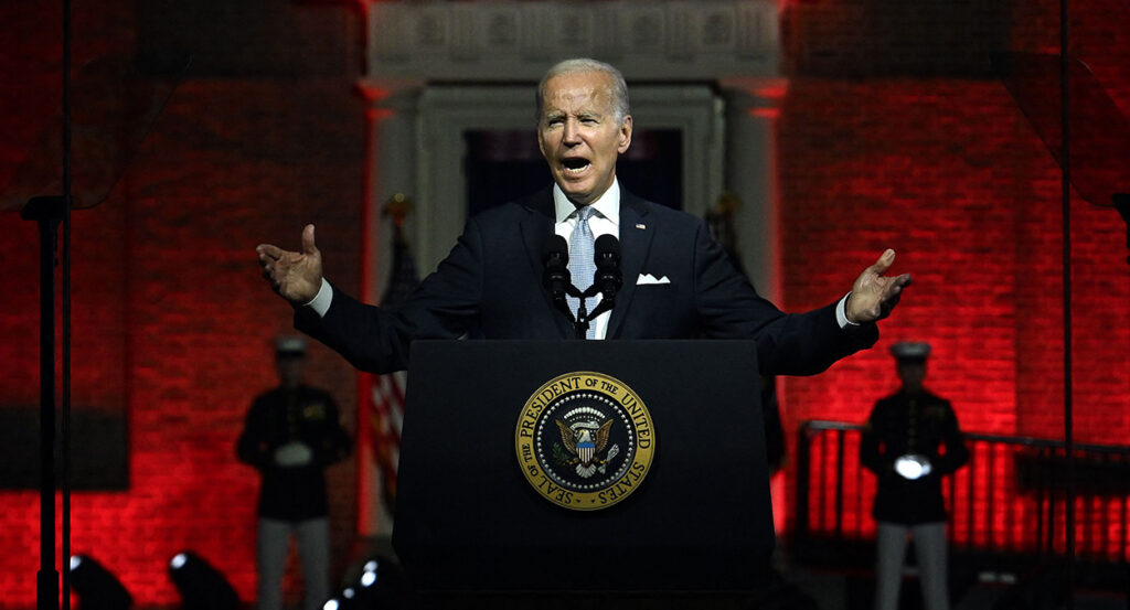 President Joe Biden in a black suit stands in front of a red-backlit Independence Hall behind a podium with the presidential seal.