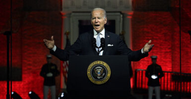 President Joe Biden in a black suit stands in front of a red-backlit Independence Hall behind a podium with the presidential seal.