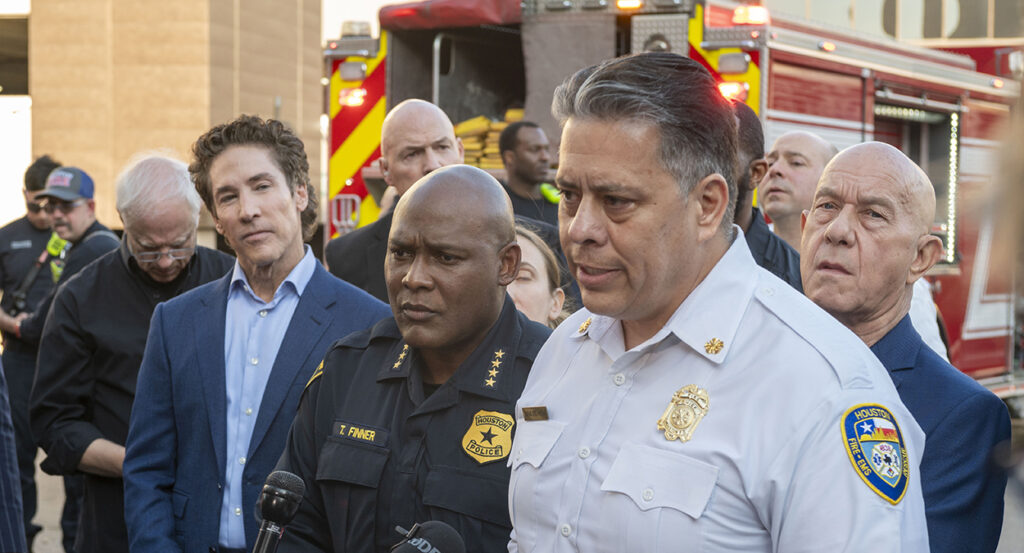 Joel Osteen in a blue suit listens as Fire Department Chief Samuel Pena and Mayor John Whitmire speak at a press conference