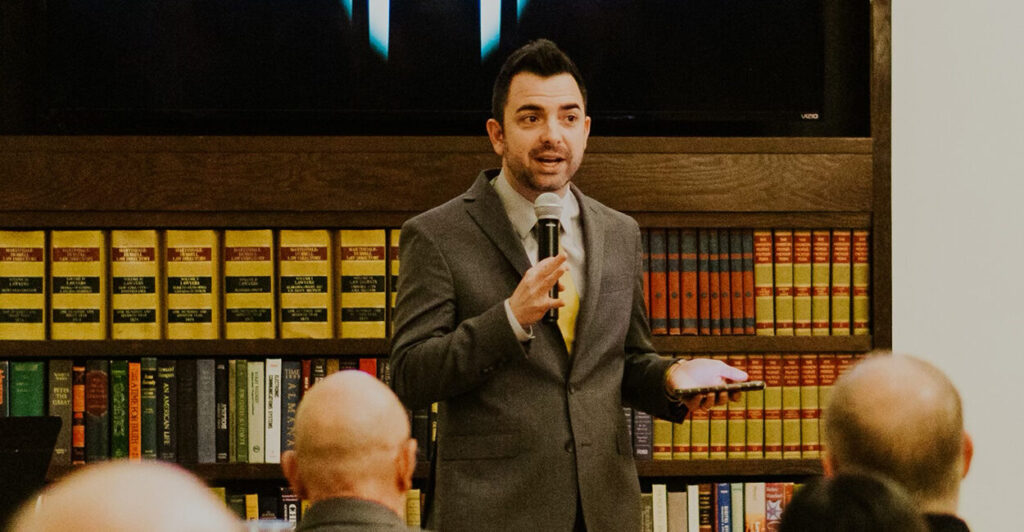 Lucas Miles in a gray suit speaks in front of bookshelves while holding a microphone