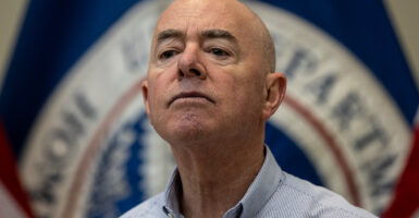Homeland Security Secretary Alejandro Mayorkas look ahead in a light blue button down shirt during a press conference with the DHS flag behind him.