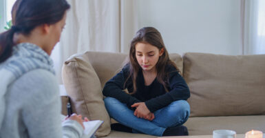 Preteen girl in jeans and a sweater crosses her hands on a couch as a therapist asks her questions with a pad and paper.