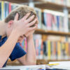 A teenage boy sits at a table, hands grabbing his hair in frustration. He is looking at a book.