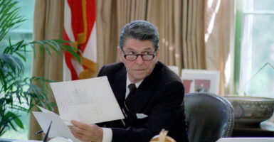 President Reagen sits at the resolute desk filled with papers. He holds up a document looking at someone off camera.