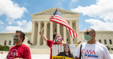 A woman waves a flag in front of the US Supreme Court Building with two people at her side holding signs and wearing shirts that say "CASA."