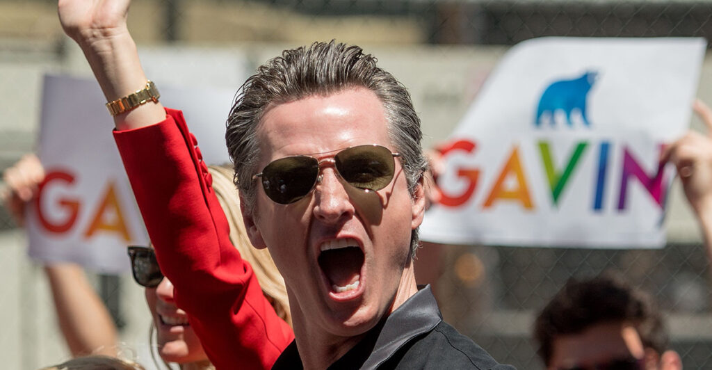 Gavin Newsom emotes with mouth open. People hold signs in rainbow font saying "Gavin."