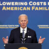 President Joe Biden delivers a speech behind a black podium Words behind him read "lowering costs for American families"