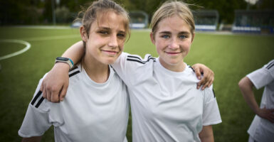 Two young girls smile for a picture, in white soccer jerseys while on a green field.