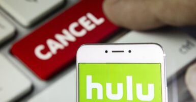 The green and white Hulu logo is seen on a phone screen, while a finger touches a red button in the back that says 