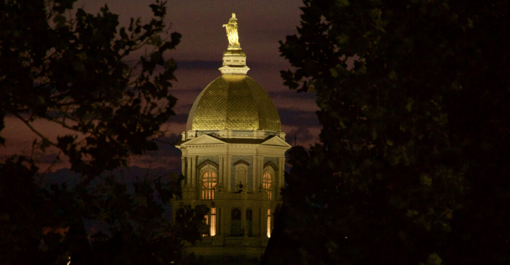 The main building of Notre Dame is lit up at night, with dark trees surrounding the illuminated tower.