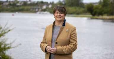 Baroness Arlene Foster stands and smile at the camera in a tan coat with a body of water behind her.