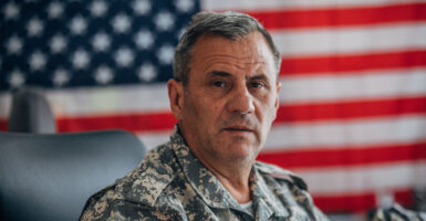 A gray-haired soldier looks at the camera while in uniform, with a United States flag behind him.