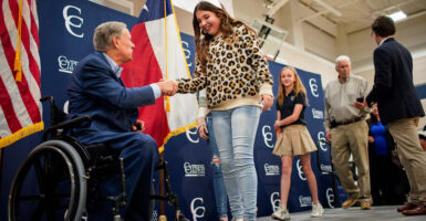 Governor Greg Abbott wears a navy blue suit, sitting in his wheelchair, while he shakes the hand of a teenaged girl wearing a cheetah print sweater.