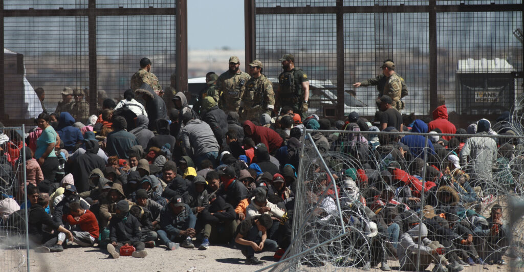 Immigrants sit in a large group by the brown border.