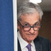 Jerome Powell, an elderly man with grey hair and facial wrinkles, looks down from his glass