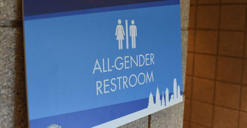 A dark and light blue bathroom sign says "ALL-GENDER RESTROOM" with two white man and woman images, side-by-side.