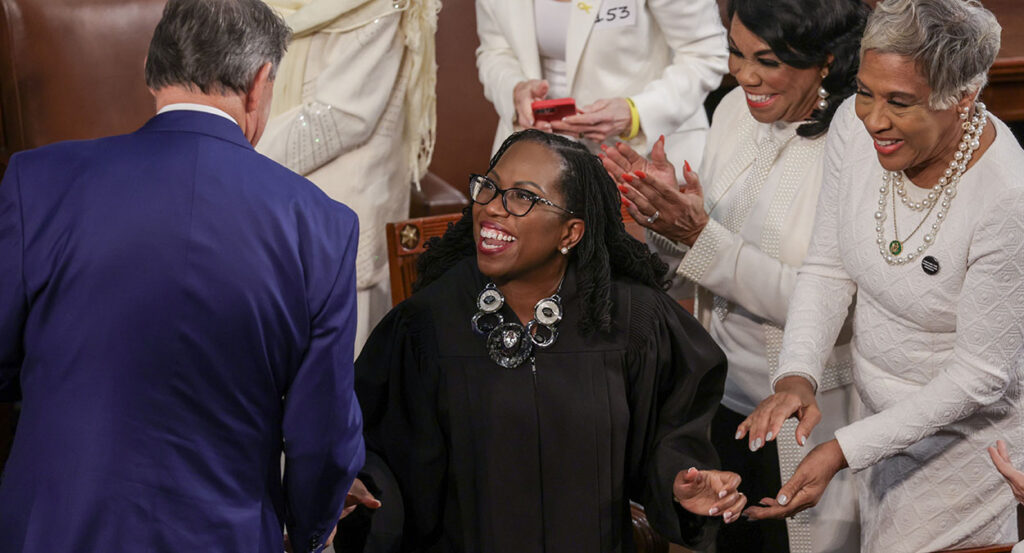 Ketanji Brown Jackson shakes hands with a man in a blue suit while she wears a large necklace above her black robes