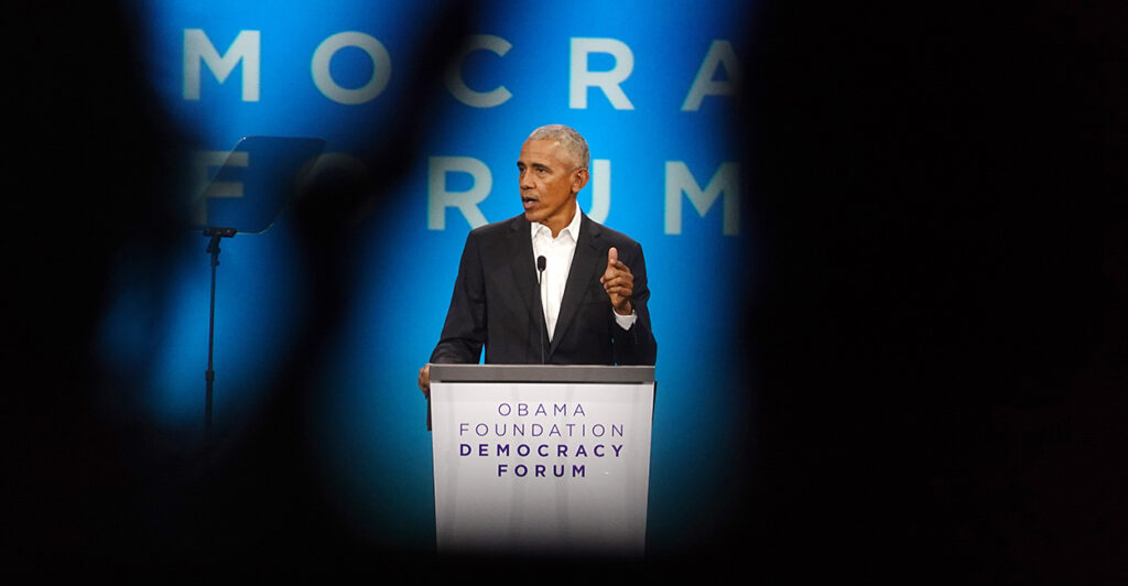 Former President Barack Obama stands at a podium and speaks in a black suit and white shirt without a tie.