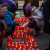 People in winter coats lights small candles in red votives on the sidewalk.
