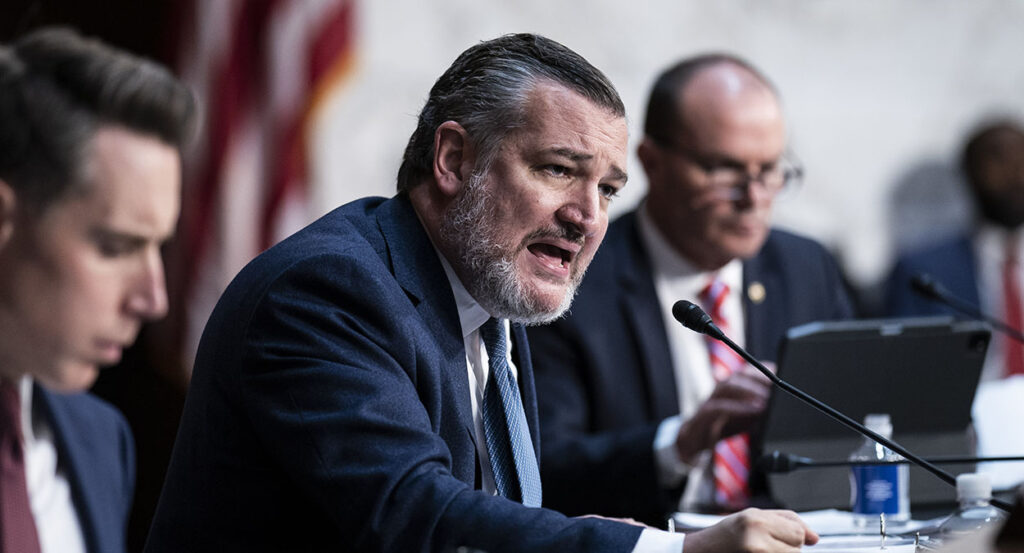 Sen. Ted Cruz, in a blue suit, pounds his fist on the table and speaks out