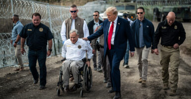Gov Abbott in his wheel chair and Trump walking travel up a dirt hill with various men behind them. A metal fence with barbed wire is next to them.