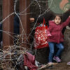small child and women immigrants walking away from steel southern border wall and barbed wire