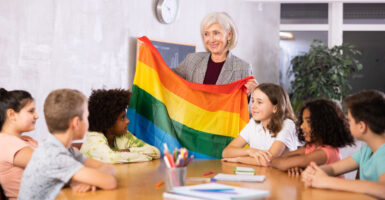 A white-haired teacher holds up a rainbow flag in front of smiling, elementary-aged children.