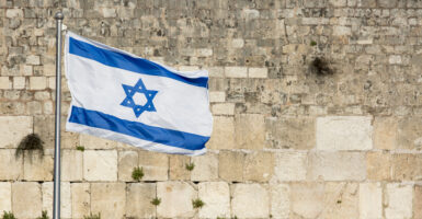 The white and blue flag of Israel flies in front of the stone Western wall.