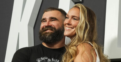 Football star Jason Kelce and his wife, Kylie, smile for a camera. Jason is wearing a black shirt, which is contrasted by Kylie's blonde hair.