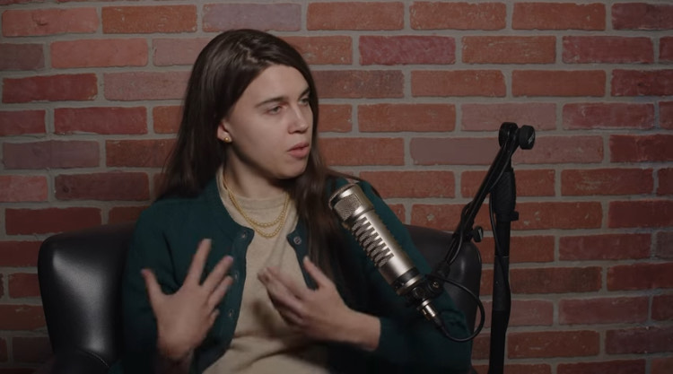 Detransitioner Camille Kiefel with her brown hair sits in front of a black microphone, while wearing a tan shirt and green sweater.