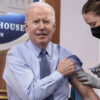 President Joe Biden receives a COVID-19 vaccine shot from a nurse while sitting down with his suit jacket off.