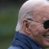 President Joe Biden smiles in a navy jacket or vest and Aviator sunglasses.