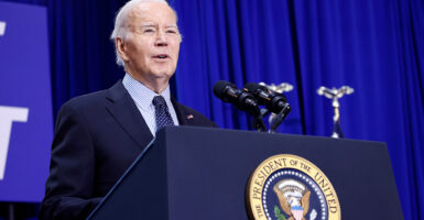 President Biden speaks at a podium in a dark suit with a royal blue curtain behind him.