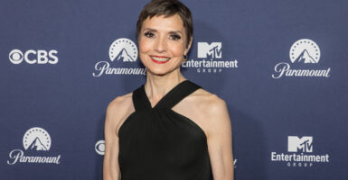 Catherine Herridge stand in front of a step and repeat in a black dress and smiles.