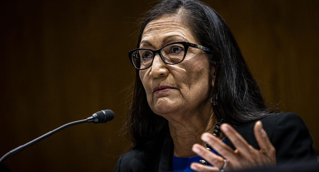 Interior Secretary Deb Haaland looks on, gesturing in front of a microphone and wearing a blue dress with a black suit jacket and pearls.