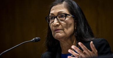 Interior Secretary Deb Haaland looks on, gesturing in front of a microphone and wearing a blue dress with a black suit jacket and pearls.