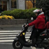 A man is seen in a red coat on a motorbike with a food delivery backpack.