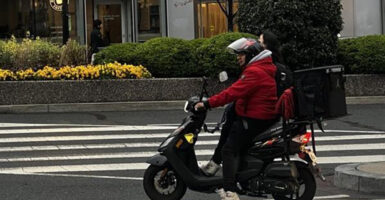 A man is seen in a red coat on a motorbike with a food delivery backpack.