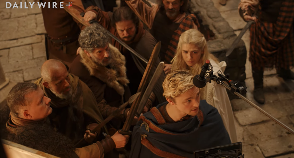 Men in medieval outfits point swords at a blond man who speaks in front of a microphone as a blonde woman stands behind him.