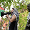 Male pro-Palestinian protester with Palestinian flag, headdress, and a megaphone alongside a bronze statue of Benjamin Franklin