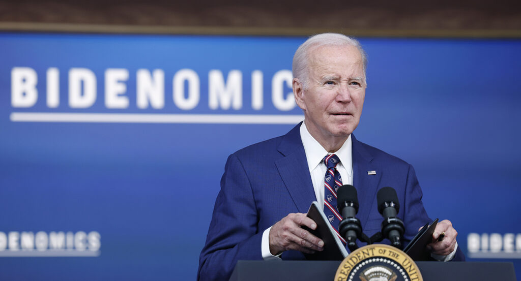 Joe Biden in a blue suit holds a briefing book behind the presidential seal and in front of a sign reading "Bidenomics."