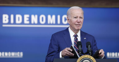 Joe Biden in a blue suit holds a briefing book behind the presidential seal and in front of a sign reading 
