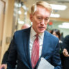 James Lankford in a blue suit looks down, holding paper and walking in a hallway.
