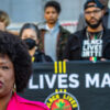 Black Lives Matter co-founder Patrisse Cullors in a pink suit stands in front of a Black Lives Matter banner with several people standing behind her on the steps of the Los Angeles City Hall.