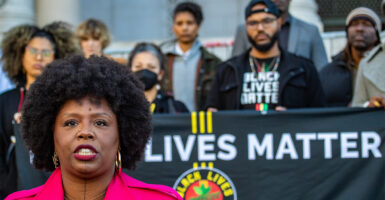Black Lives Matter co-founder Patrisse Cullors in a pink suit stands in front of a Black Lives Matter banner with several people standing behind her on the steps of the Los Angeles City Hall.